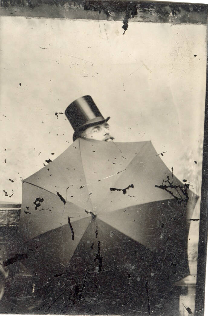 Black and white photograph of a man in a top hat, peaking over the edge of an opened umbrella.